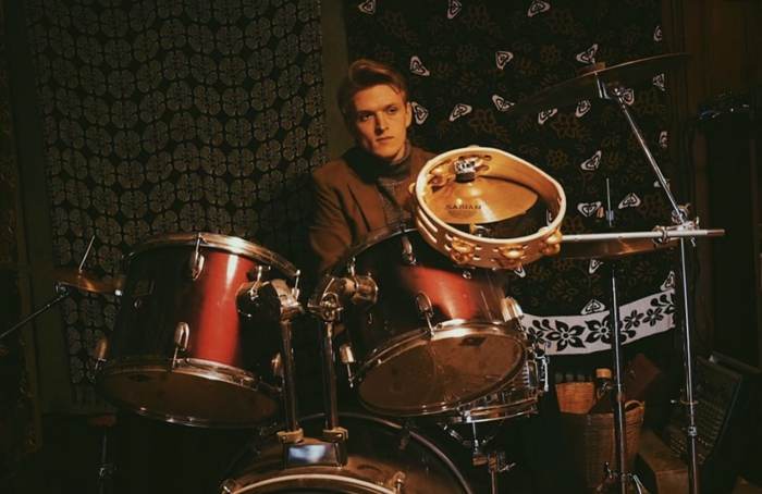 Behind a drum kit with tapestries on the wall